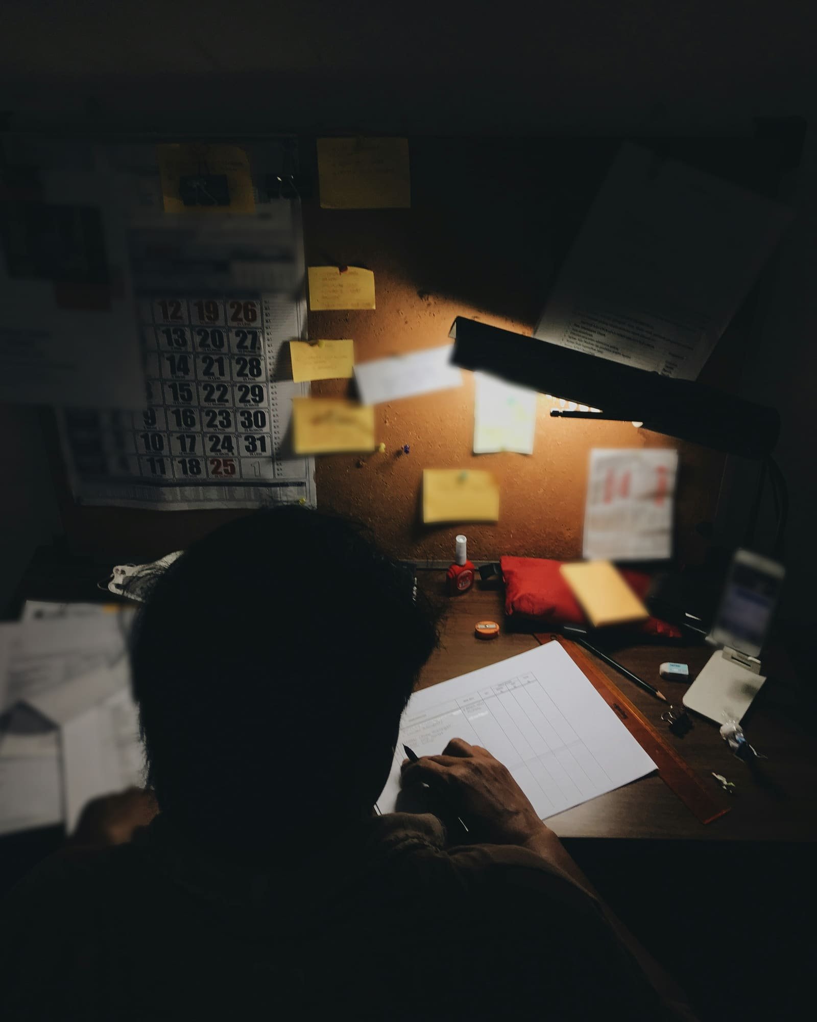 Late at night, a person seen from behind fills in a ruled ledger sheet under a desk lamp, a wall calendar and sticky notes pinned to a corkboard above