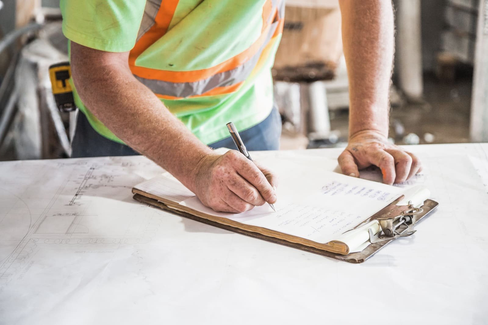 A worker in a high-visibility shirt writing on a clipboard laid over drawings, face out of frame