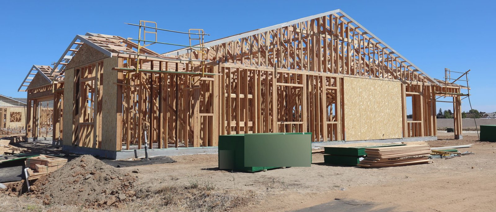Wood framing of new single-storey houses on a dirt lot under a clear sky, bundles of sheathing and lumber on the ground