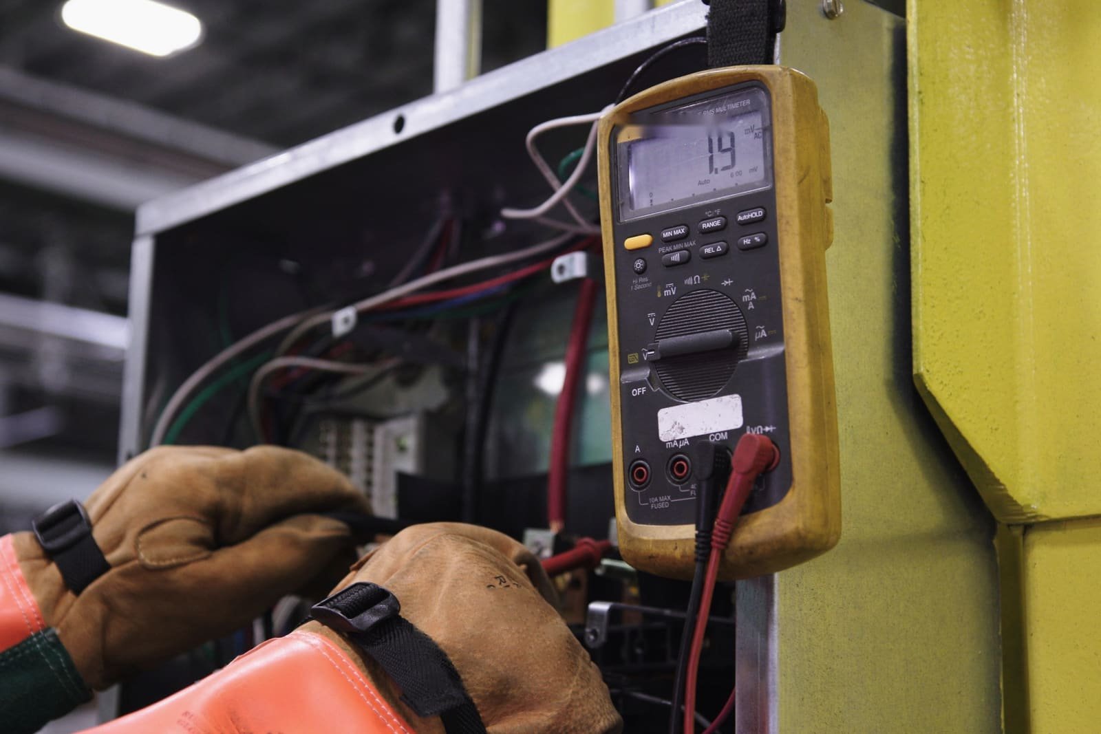 Gloved hands working inside an open electrical panel, a multimeter clipped beside it
