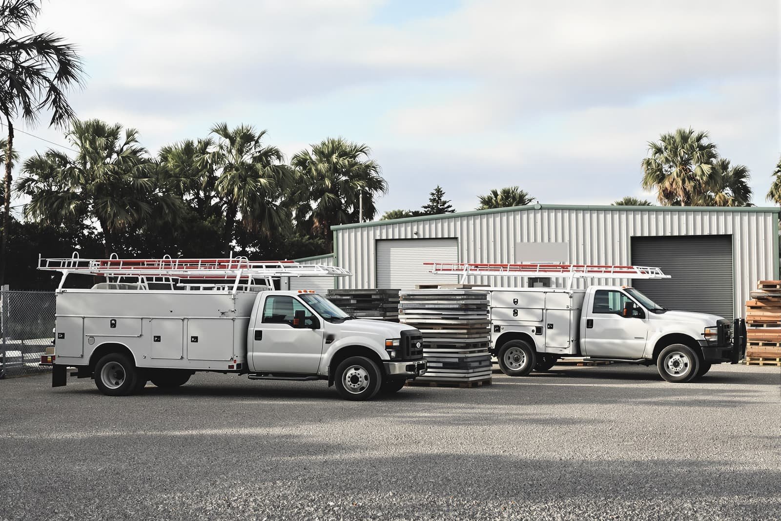 Two white service trucks with ladder racks parked on gravel outside a metal building, palms behind