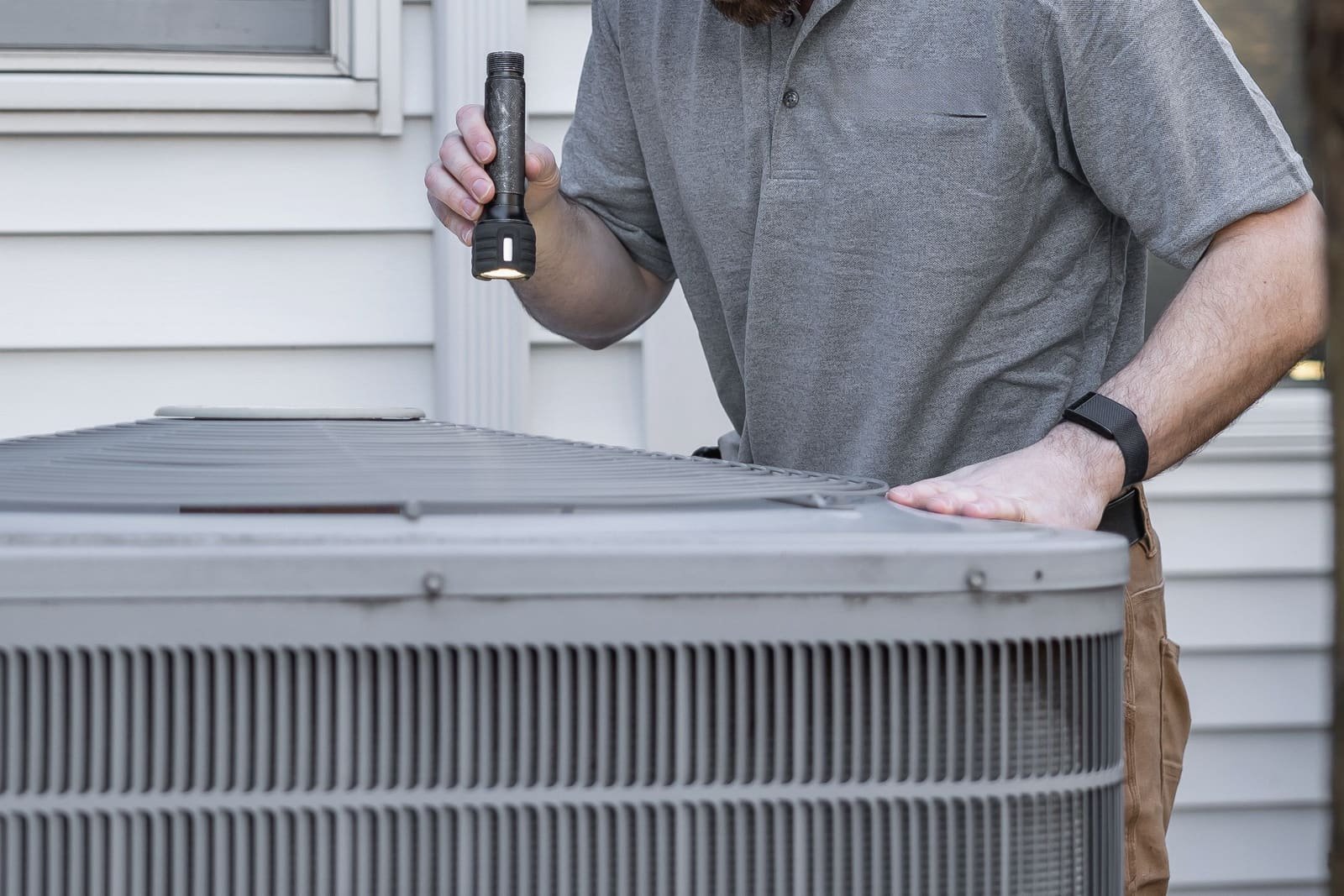 A technician with a flashlight checking an outdoor air-conditioning condenser beside a house, face out of frame