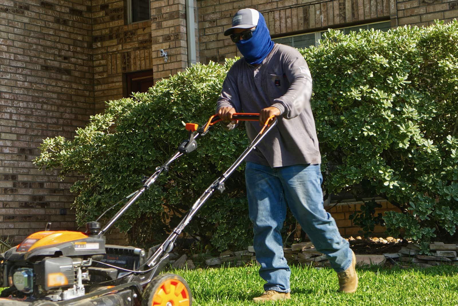 A lawn-care worker in a sun mask and cap pushing a mower across a front lawn beside a brick house