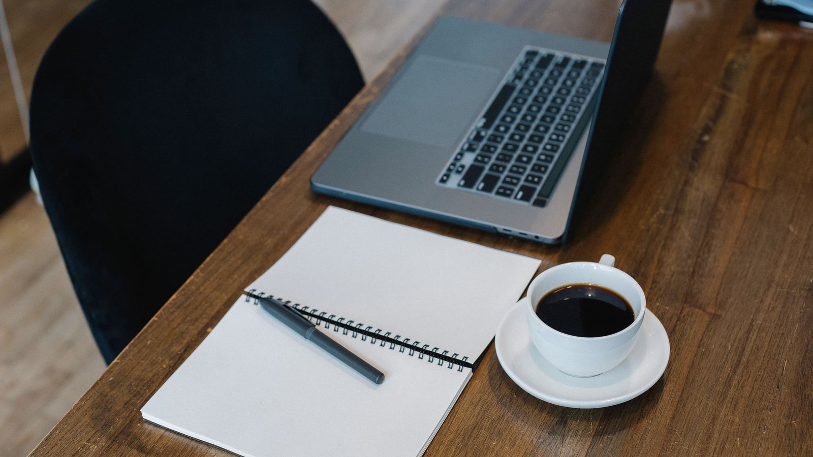 A wooden desk with an open laptop, a blank spiral notebook and pen, and a cup of black coffee